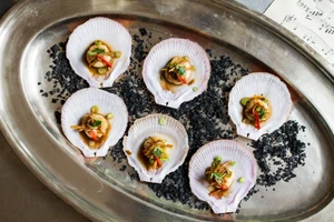 Top-down view of six scallops in half shells on a silver platter with black flakes and a sheet of paper.
