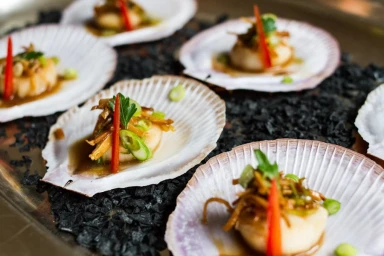 Seared scallops served on individual white shells over dark charcoal, garnished with red chili strips and green onions.