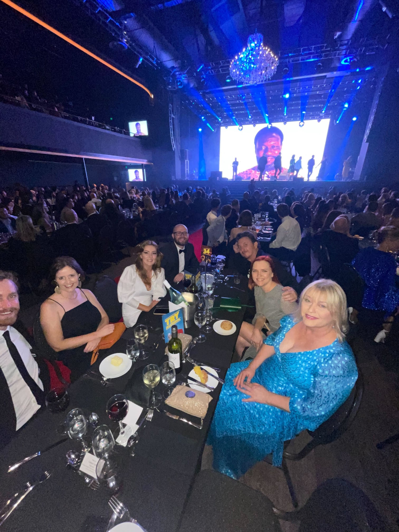 Indoor event with a large group of people seated at tables in a dark event space, focused on a stage with a performer visible on screen.