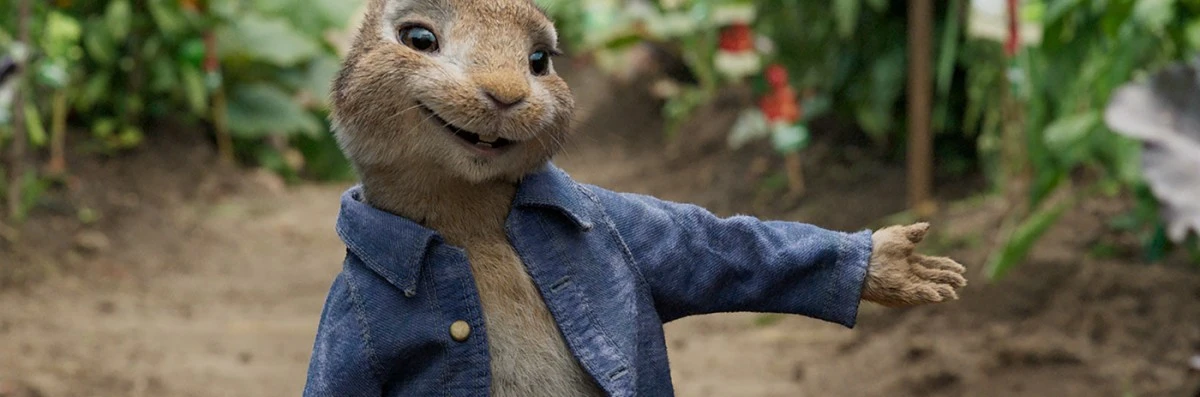 Anthropomorphic rabbit character in a blue denim jacket smiles with an open palm in an outdoor garden setting with blurred greenery.