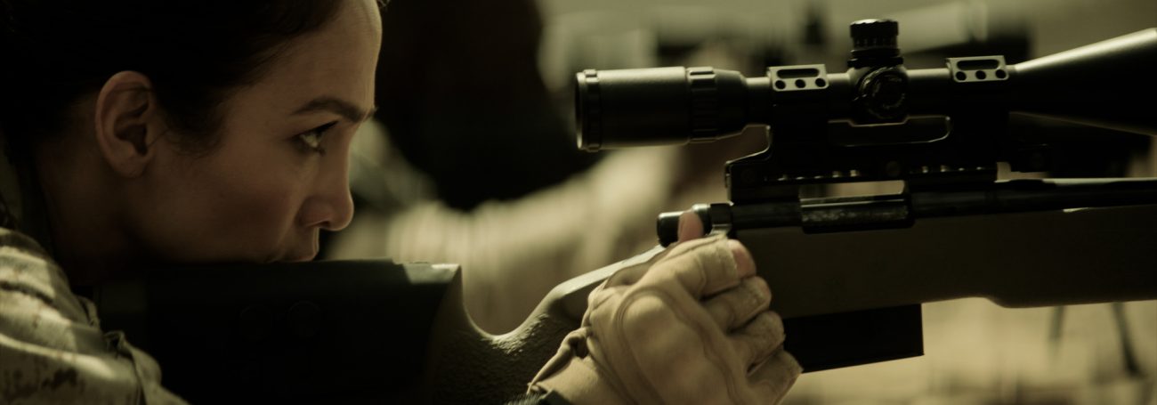 Adult woman aiming a sniper rifle with scope, close-up on her face and hands, focused on the weapon, indoor environment with dark lighting.