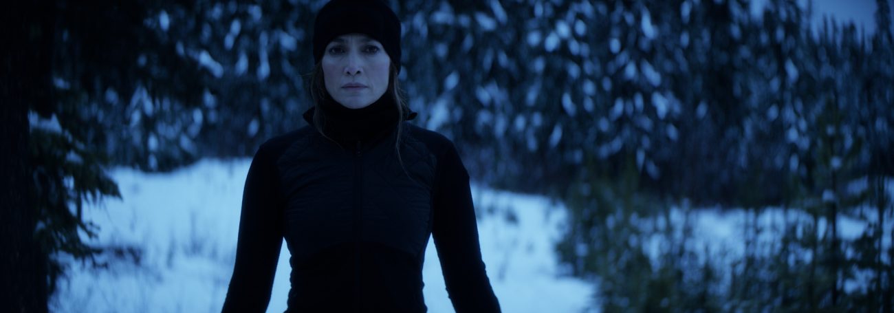 Adult female in black beanie and turtleneck, gazing forward in snowy forest landscape, with blurred pine trees in soft, dim lighting.