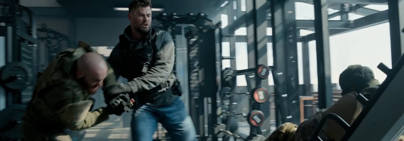 Action shot of two adult males engaged in physical combat in a gym setting with weightlifting equipment and neutral color palette.