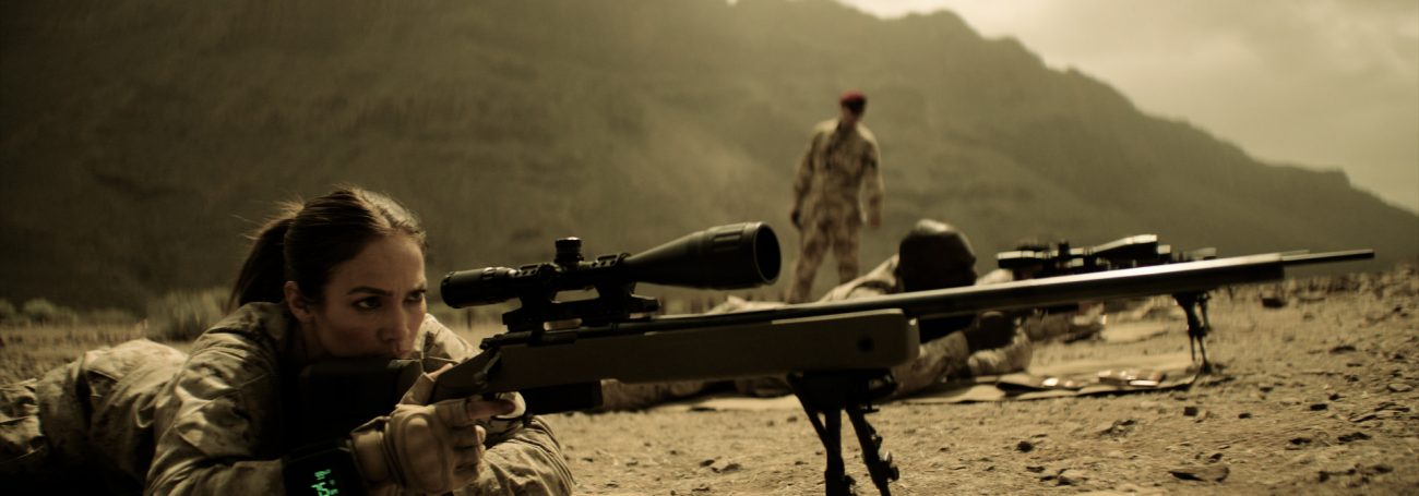 Female sniper aims rifle with scope outdoors in desert landscape, wearing camouflage uniform with second soldier and mountains visible.