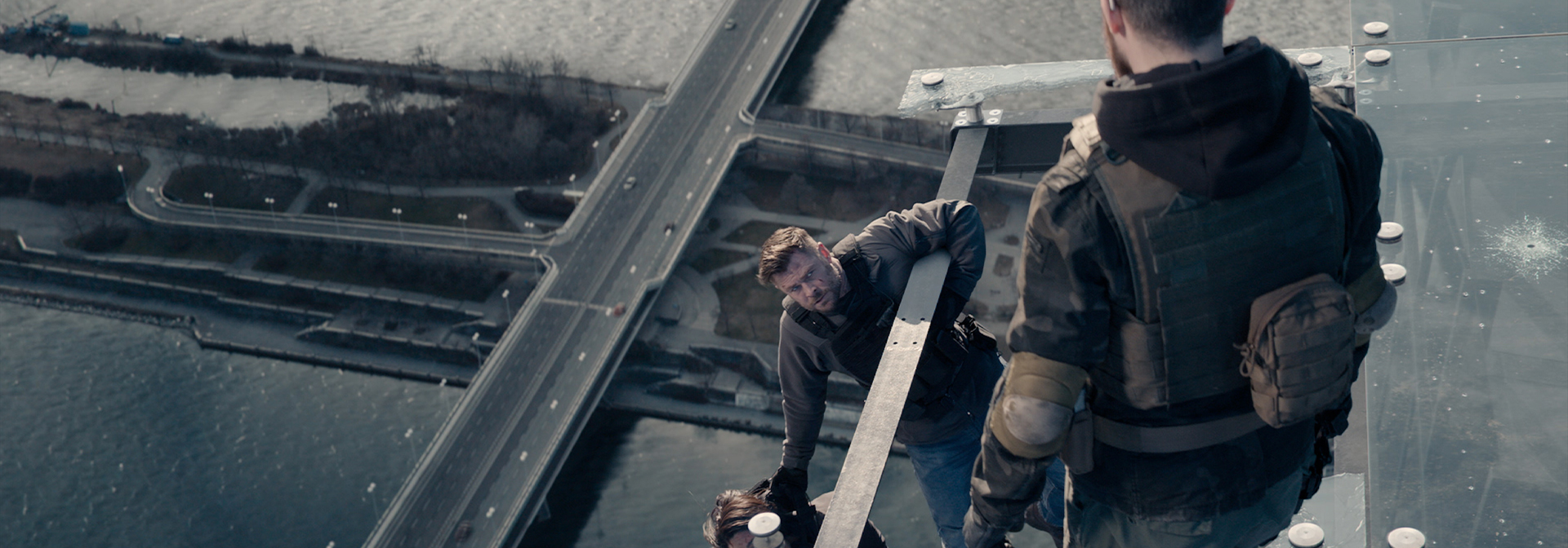 Action scene of two men on a glass rooftop with a cityscape in the background; one man is helping another in a moment of tension.