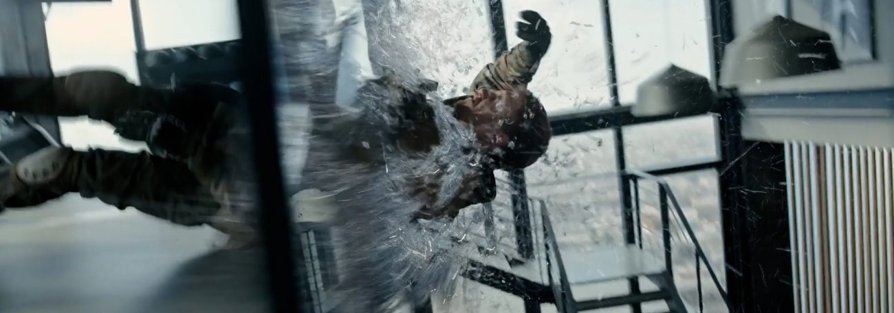 Man falling backwards through a glass pane indoors, wearing a khaki jacket and dark pants. Staircase, interior, and window visible.