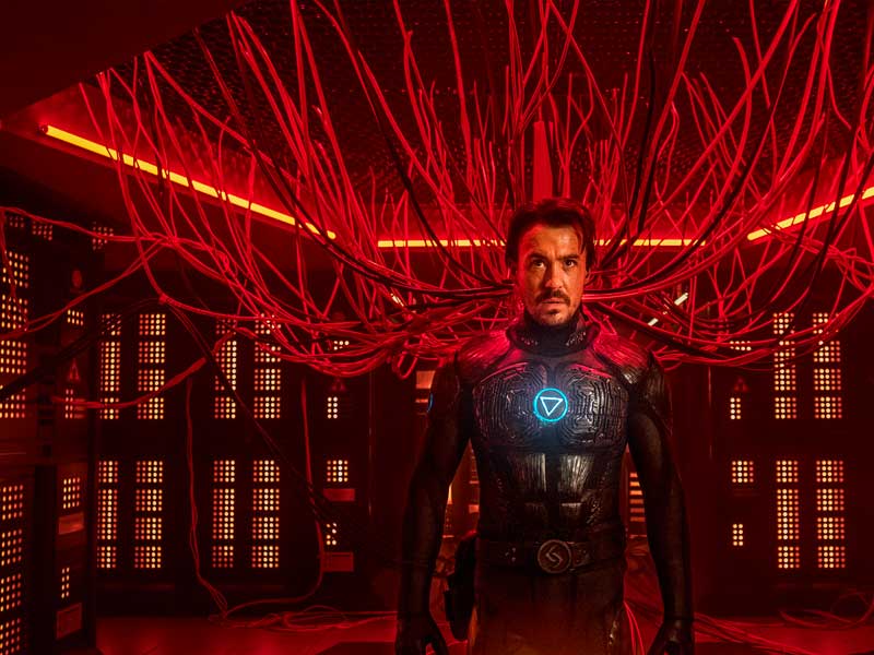 Man in dark futuristic suit with glowing emblem stands amidst a network of red cables and computer equipment in a dimly lit interior.