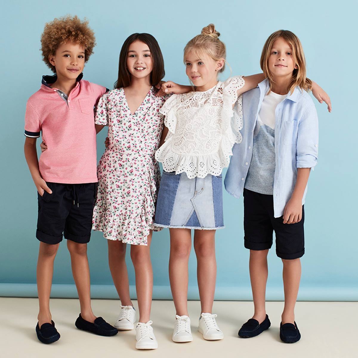 Four smiling children, two boys and two girls, stand together in a studio with a blue background, showcasing kids' fashion.