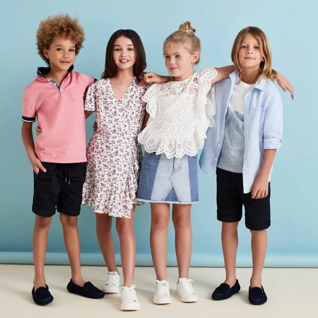 Four smiling children, two boys and two girls, stand together in a studio with a blue background, showcasing kids' fashion.