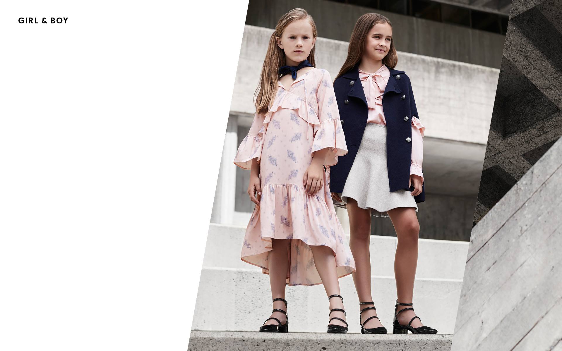 Two young girls in fashionable outfits pose on concrete steps. One wears a pink dress, the other a navy coat and skirt in an urban setting.