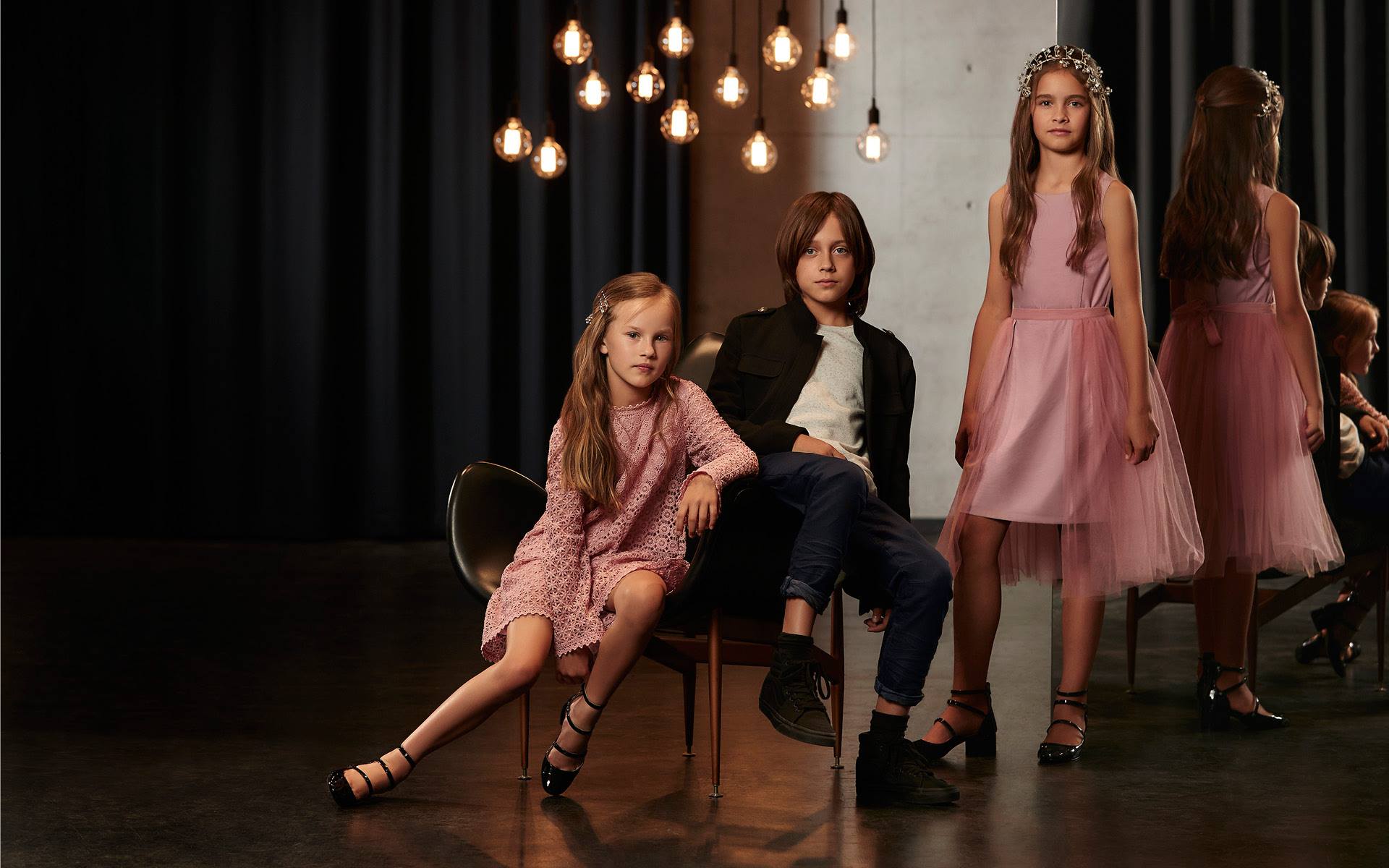 Group of three young girls and one boy posing in pink dresses and casual clothes with dark background and hanging light bulbs