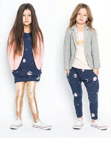 Two young girls model children's fashion, wearing patterned tops and bottoms, blazers, leggings, and Converse shoes in a bright photo studio.