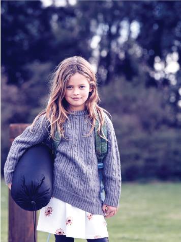 Young girl in gray sweater and flower skirt holds sleeping bag outdoors on a blurred backdrop of trees and a cloudy sky.