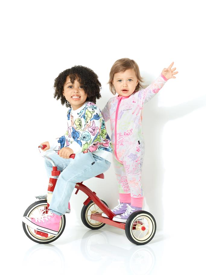 Two young children, a boy on a red tricycle and a girl standing, both smiling in a light and airy indoor studio setting.