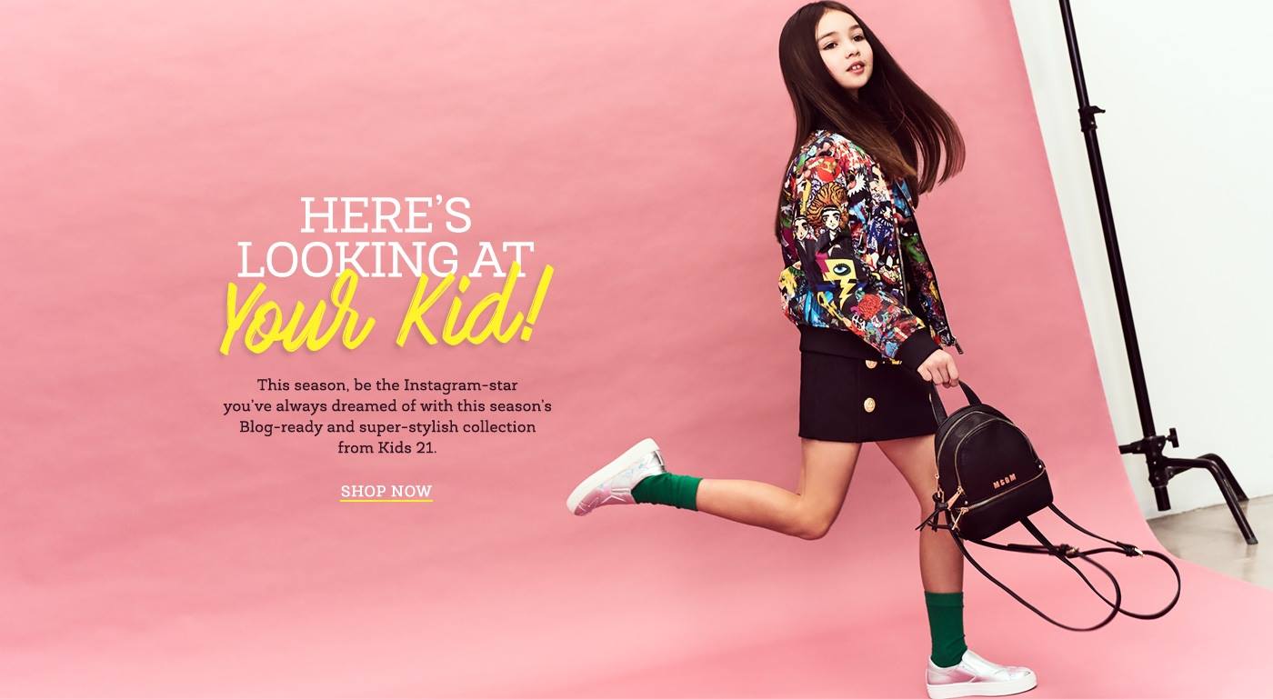 Full-length shot of a child model wearing a patterned jacket, black skirt, green socks and white sneakers holding a backpack in a studio.