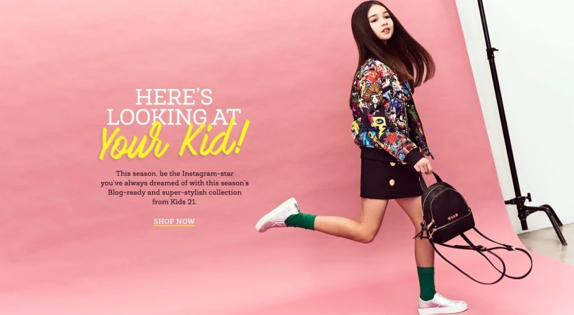 Full-length shot of a child model wearing a patterned jacket, black skirt, green socks and white sneakers holding a backpack in a studio.