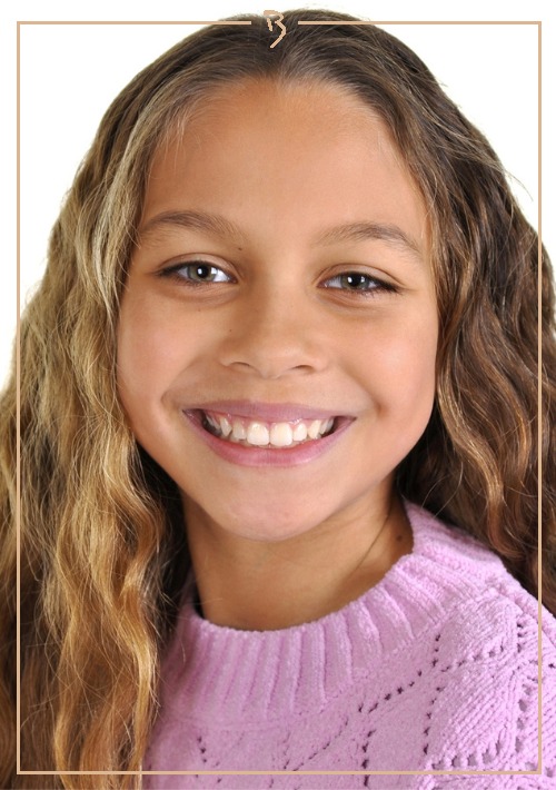 Portrait of a smiling young girl with long wavy hair wearing a lilac sweater against a white background in a framed composition.