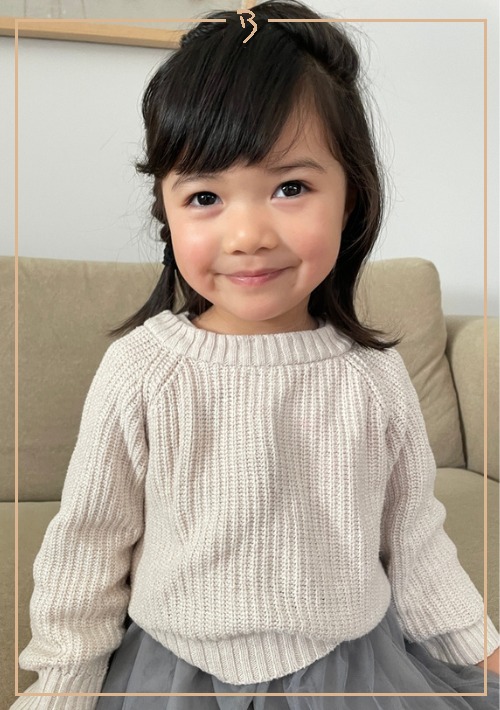 A young child with dark hair and a beige sweater smiles sweetly in a close-up studio portrait with soft, diffused lighting.