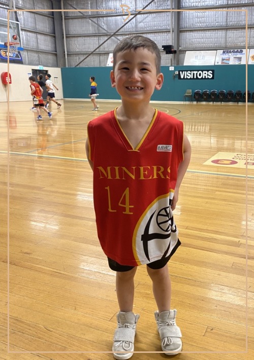 Young boy in a red and gold basketball jersey smiles indoors on a polished basketball court with other players practicing.