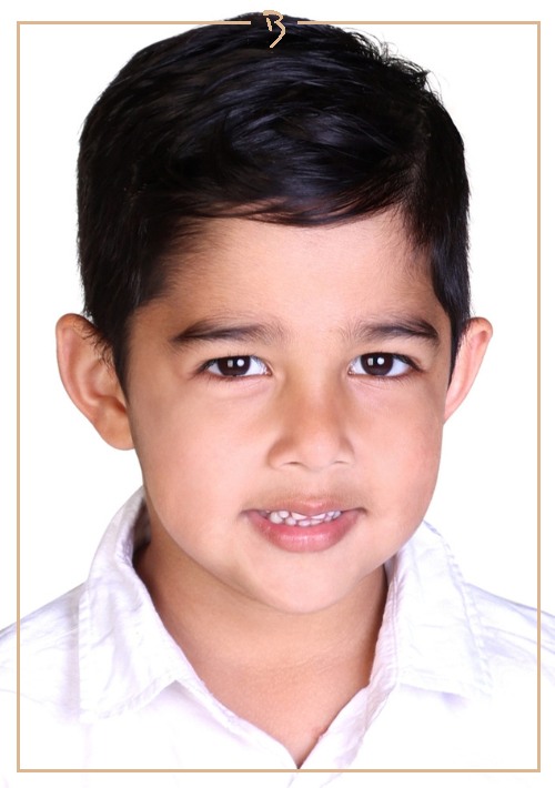 Close-up portrait of a young child smiling indoors, wearing a white collared shirt against a plain white backdrop, framed in a golden border.