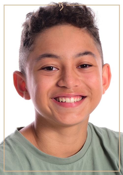 Close-up portrait of a smiling boy with curly hair, fair skin, and a green shirt against a white background and thin gold border.