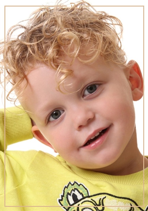 Close-up portrait of a smiling toddler with curly blonde hair, wearing a yellow shirt, against a white background in a studio shot.