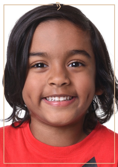 Portrait of a smiling child with dark hair and a red shirt, captured in a close-up shot against a bright white background.