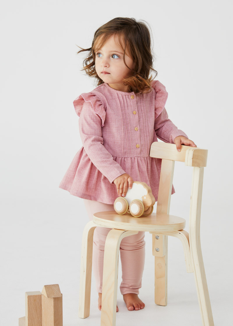 Young girl in a pink dress stands next to a light-colored wooden chair, holding a wooden toy car, with wooden blocks nearby on white.