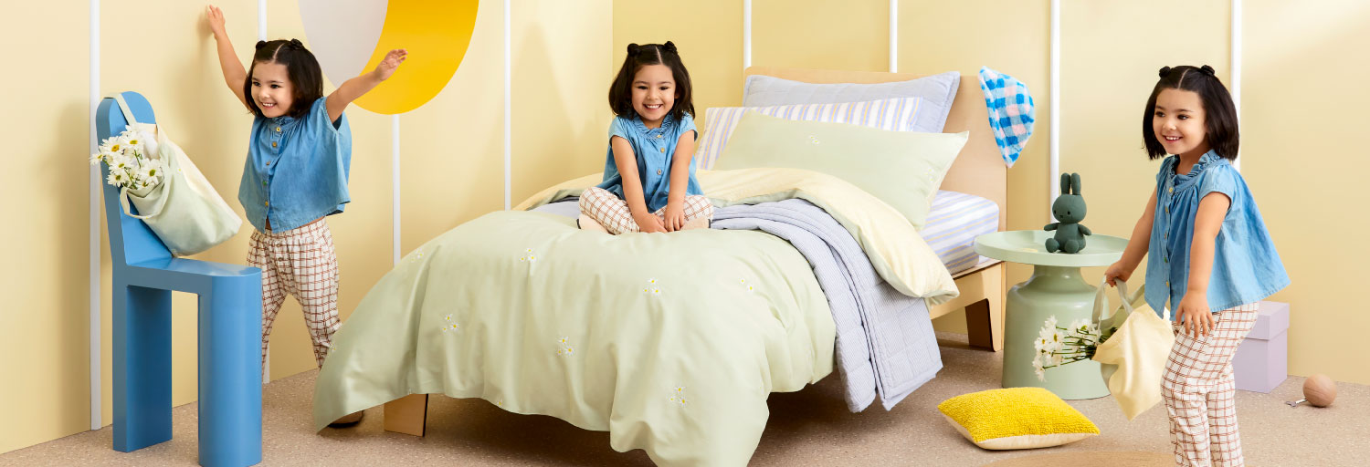 Three young girls in a bedroom setting, with a bed, chair, and decorative items, all bathed in soft, pastel colors and natural lighting.