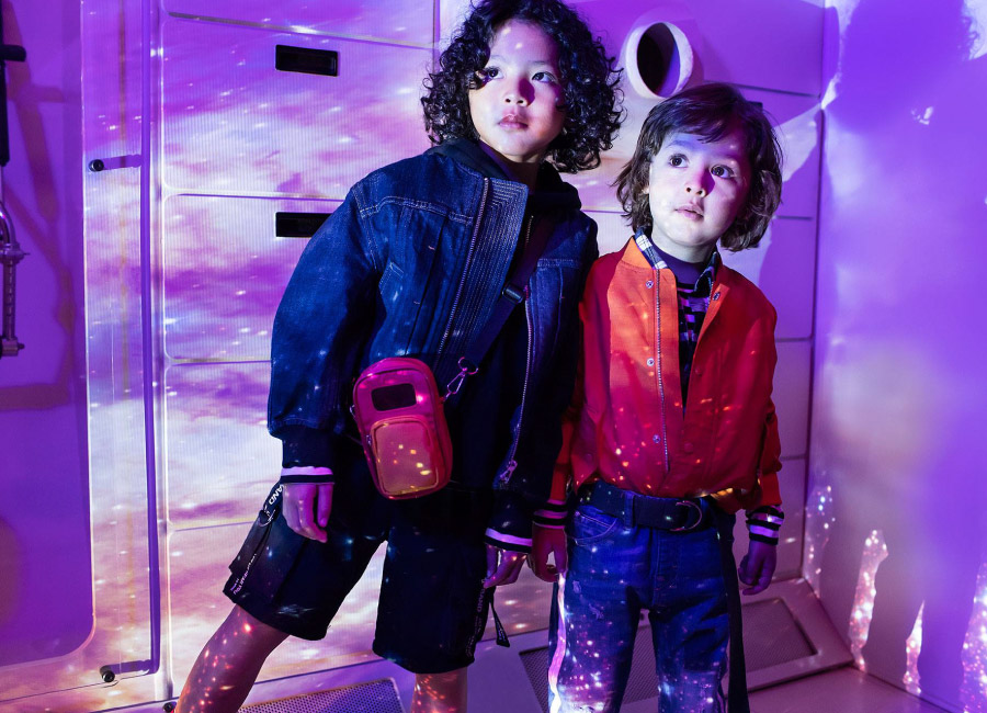 Two children model contemporary outfits with denim jacket, orange bomber jacket, and space-themed, galaxy-printed background with neon lighting.