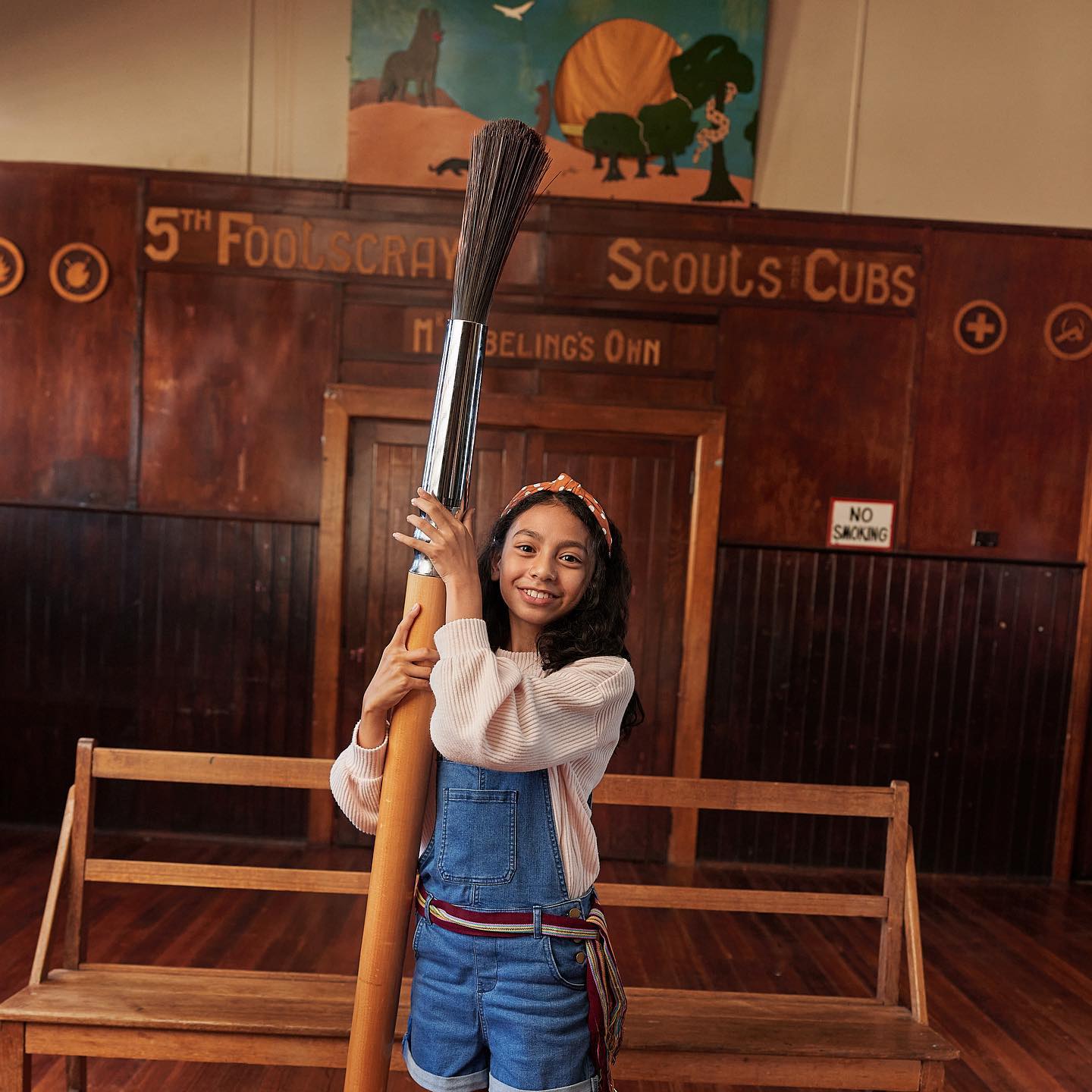 A young girl holds a very large paintbrush inside of a building with a wooden bench, panelled walls, and a sign above a doorway.
