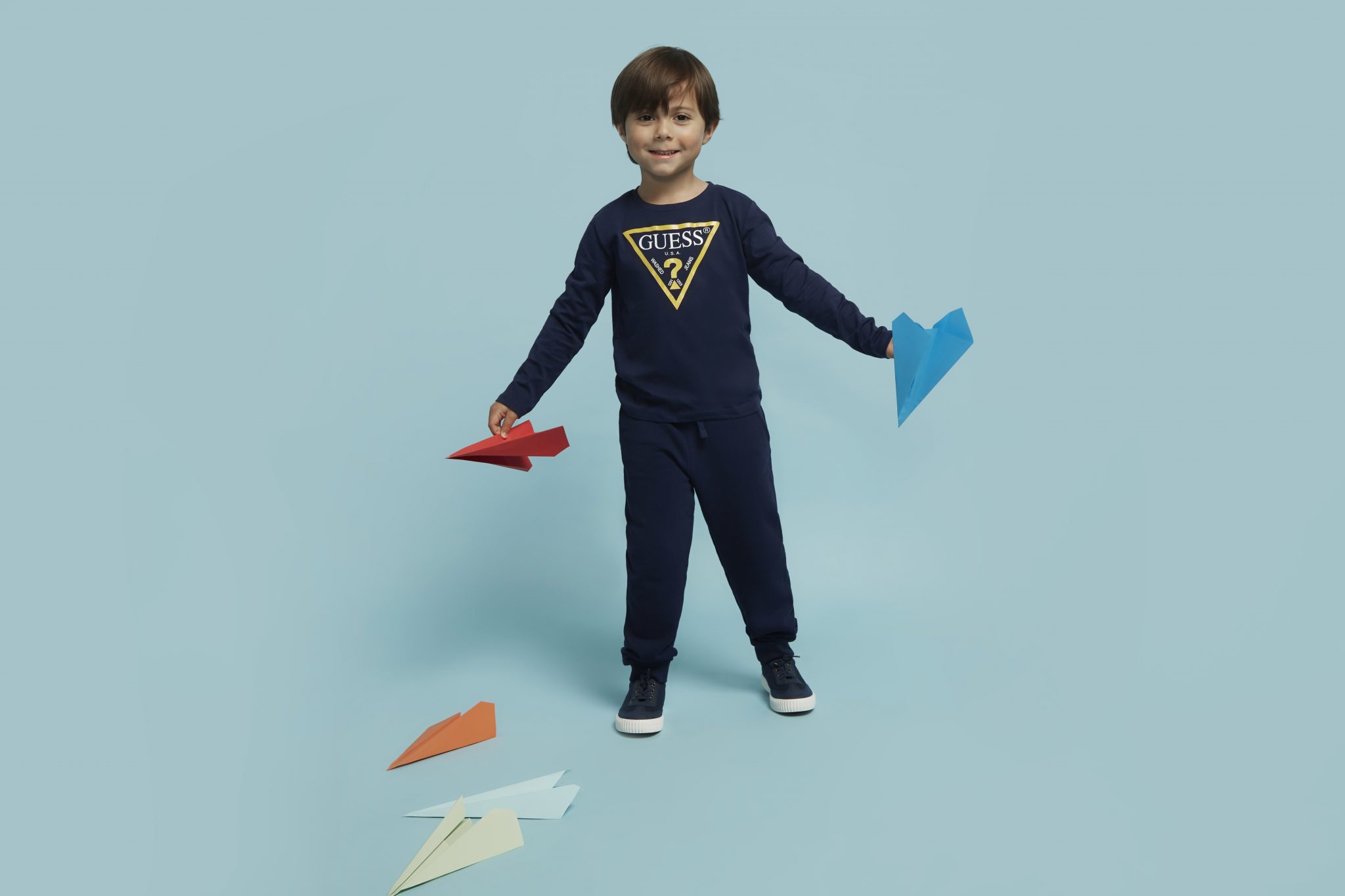 Boy poses in navy blue sweat suit on light blue backdrop, holding red and blue paper airplanes. Other paper airplanes are scattered on floor.