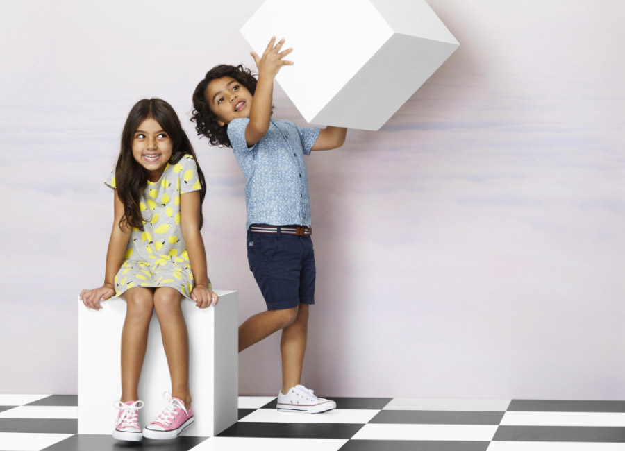 Two smiling children, a girl sitting and a boy standing, pose with a white cube against a gradient backdrop and checkerboard floor.