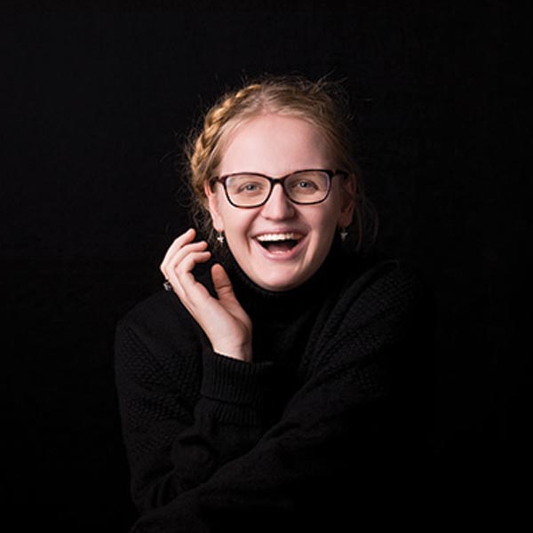 Smiling blonde woman in a black turtleneck sweater with a braided hairstyle and glasses poses in a studio with a dark background.