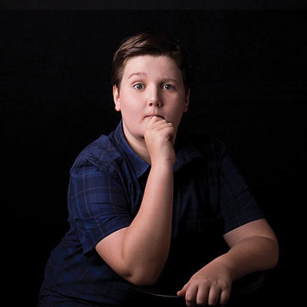 Pensive child in blue plaid shirt, with a hand to the mouth, seated against a dark backdrop in a portrait-style composition.