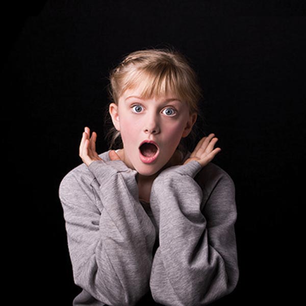 Surprised fair-skinned child with blonde braided hair and blue eyes, mouth open, hands up near face, gray sweater, dark background.