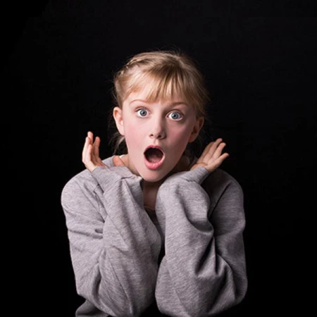 Surprised fair-skinned child with blonde braided hair and blue eyes, mouth open, hands up near face, gray sweater, dark background.