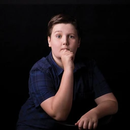 Young boy in a dark blue short-sleeve shirt with a hand near his chin against a black studio background.