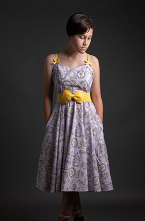 A young woman poses in a vintage-inspired light purple dress with a yellow bow belt, displayed against a dark grey studio backdrop.