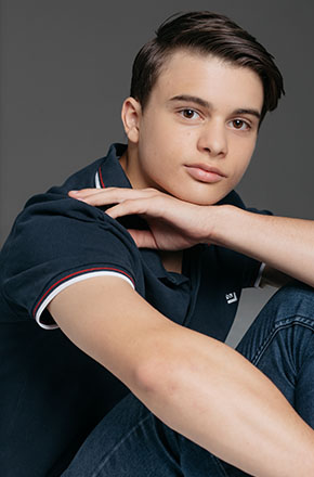 Portrait of a young man in a blue polo shirt and jeans, posing indoors against a grey backdrop, lit with soft studio lighting.