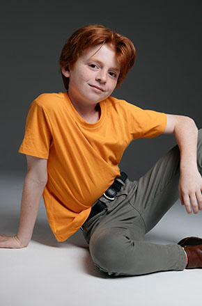 Portrait of a red-haired child with a slight smile, wearing an orange t-shirt and seated on a gray and white background in a studio.
