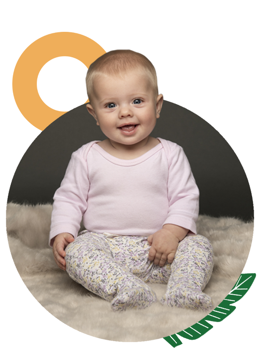 Adorable baby sits on a fluffy surface, wearing a light pink shirt and patterned pants, smiling with a black and grey graphic background.