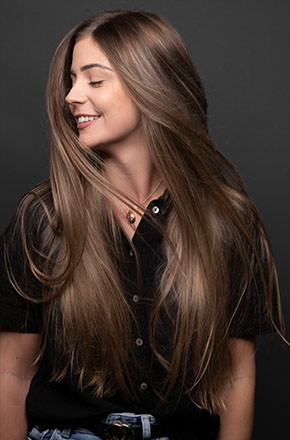Portrait of a smiling young woman with long brown hair wearing a black button-down shirt against a dark gray studio background.