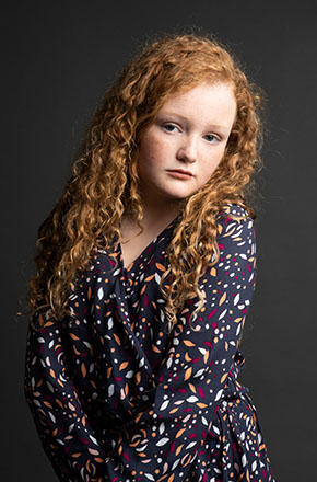 Portrait of a young woman with long red curly hair wearing a patterned wrap dress, posing in a photo studio against a dark background.