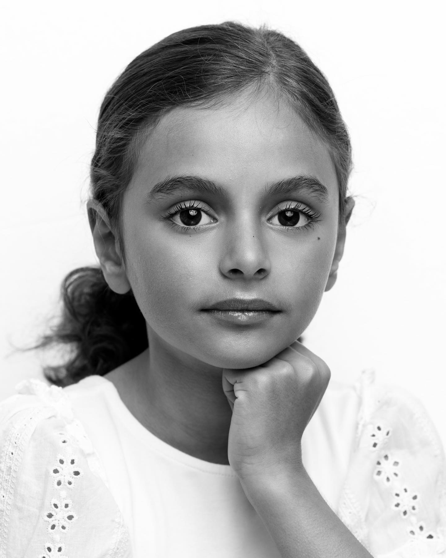 Close-up portrait of a young girl with dark hair and eyes, fair skin, wearing a white embroidered shirt against a white backdrop.