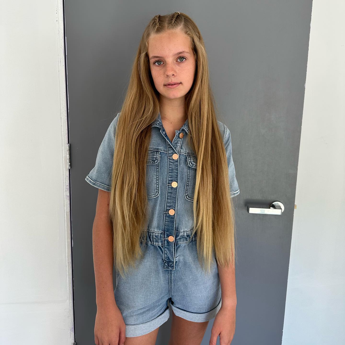 Young girl with long blond hair wears a denim romper against a gray door in an eye-level medium shot; indoor setting.