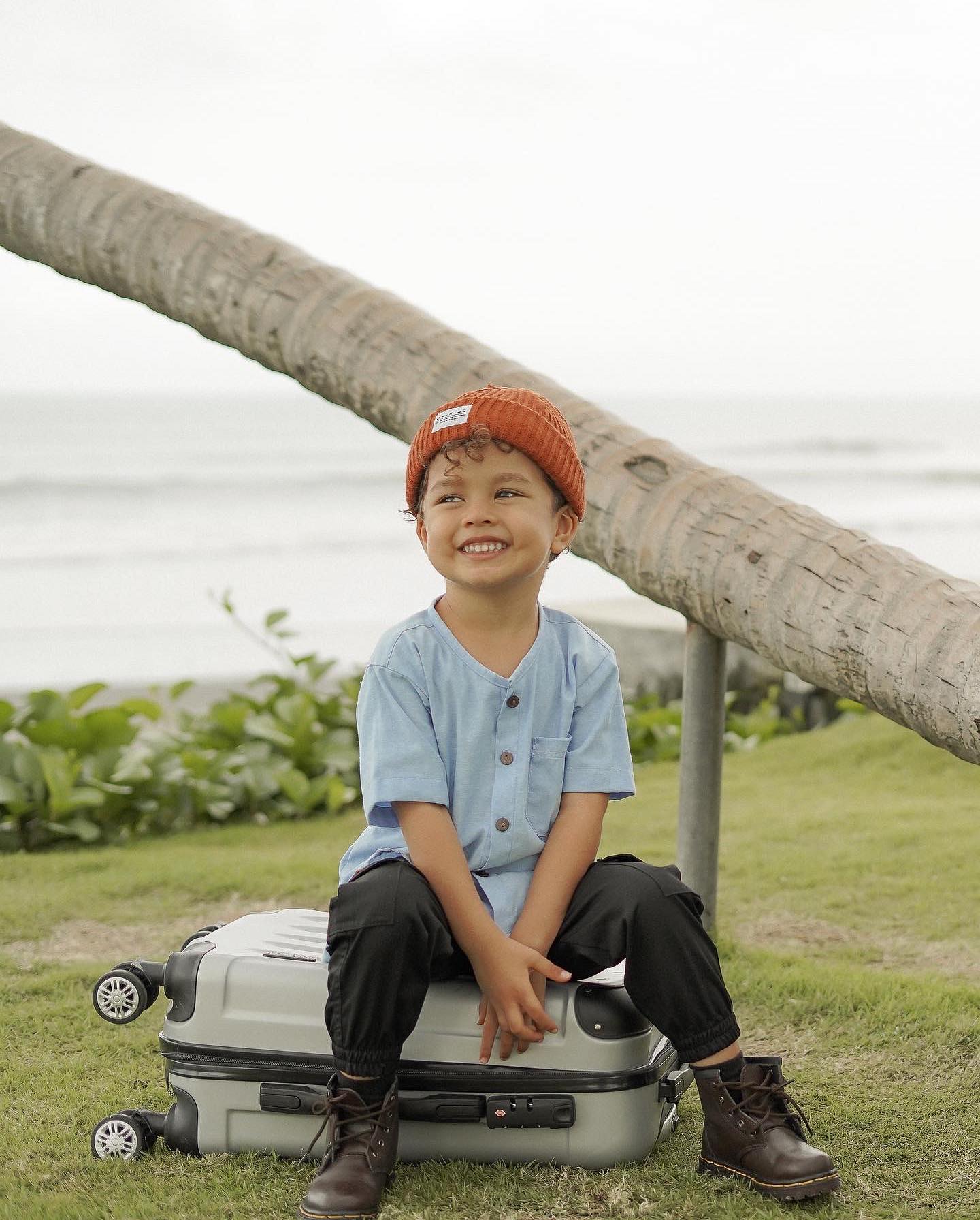 A smiling young boy in a beanie sits on a suitcase near the ocean, posing for a lifestyle portrait in natural light.