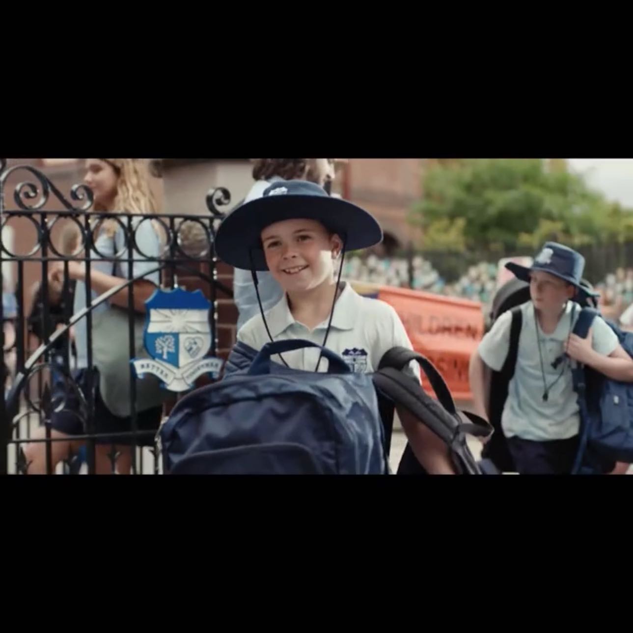 A smiling schoolboy wearing a hat and carrying a backpack walks through school gates. Other children are in the background.