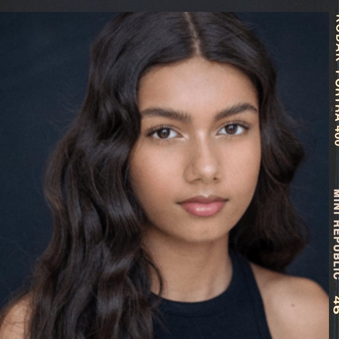 Close-up portrait of a young woman with long, wavy dark hair, fair skin, and a neutral expression against a dark background.