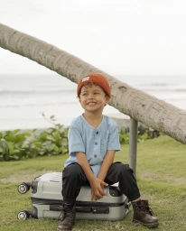 Young child wearing an orange beanie and blue shirt sitting on a silver suitcase on grass near a beach